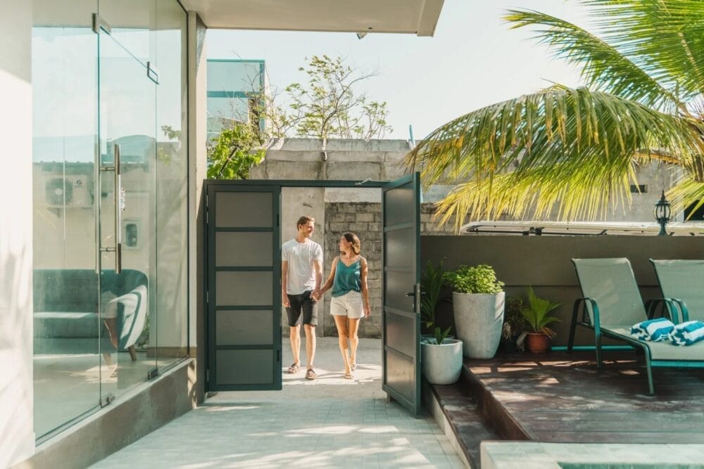 Una coppia che si tiene per mano attraversa un cancello per entrare nel moderno patio esterno dello SKY BEACH HOTEL, con sedie a sdraio, piante in vaso e una palma accanto a un edificio con pareti di vetro in una giornata di sole.