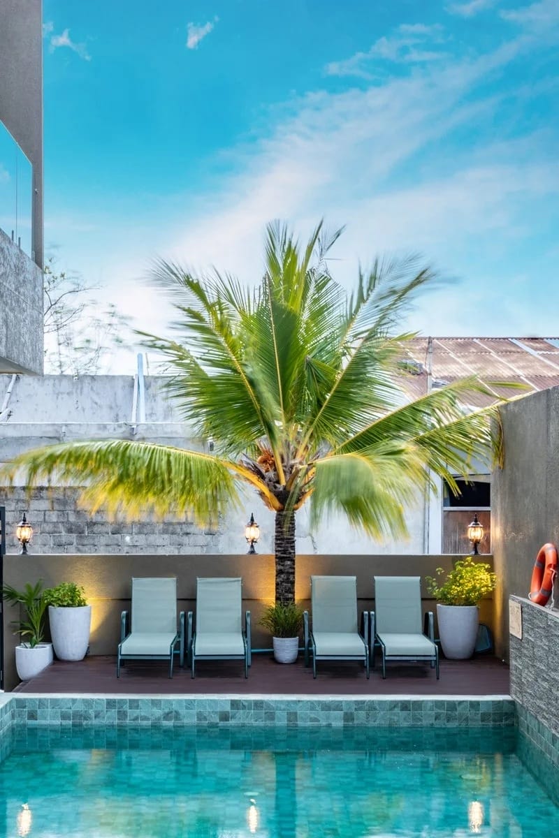 La moderna area della piscina all'aperto dello SKY BEACH HOTEL è caratterizzata da sei sedie a sdraio sotto una grande palma, affiancate da piante in vaso e lampade a muro, con l'acqua limpida in primo piano e il cielo luminoso sopra.