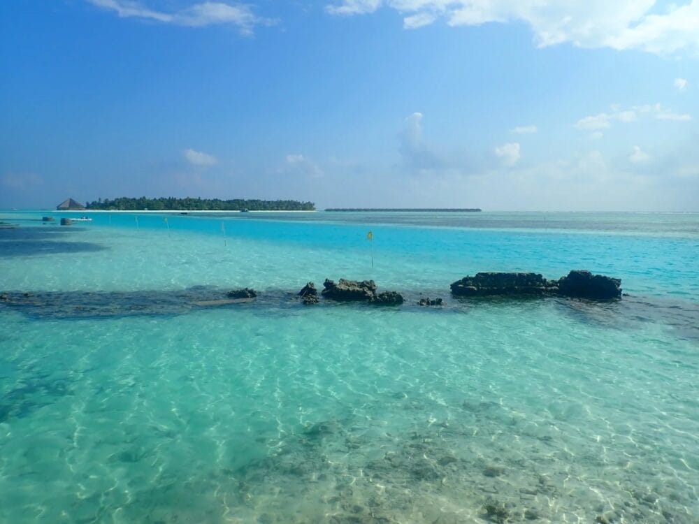 L'acqua limpida e turchese dell'oceano con le rocce scure in primo piano, un'isola lontana fiancheggiata da palme e l'invitante Sky Beach Hotel annidato sotto un cielo blu brillante con nuvole sparse sopra.