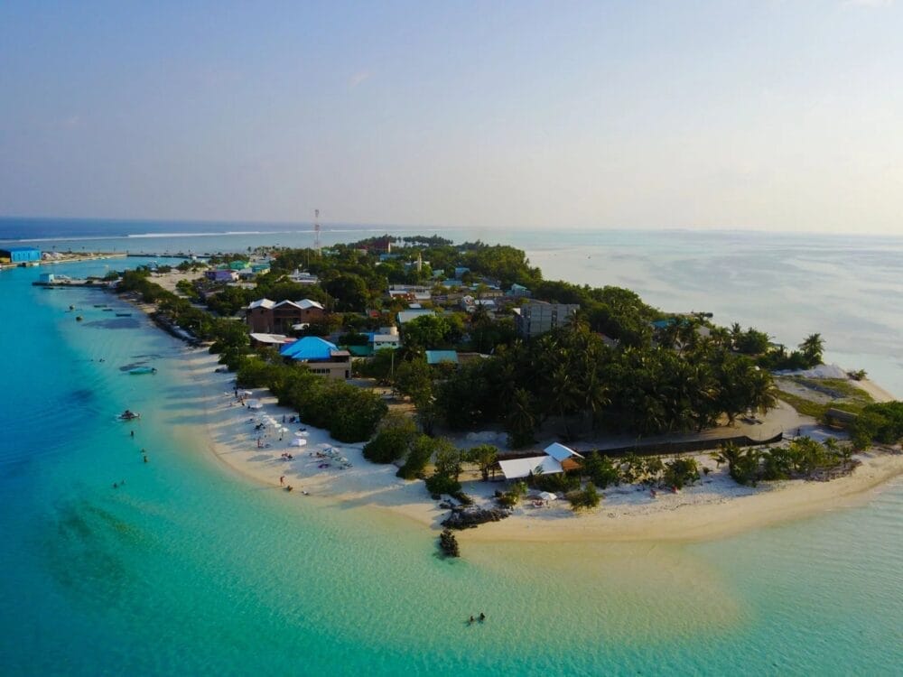 Veduta aerea di un'isola tropicale con acque turchesi, spiagge sabbiose e vegetazione lussureggiante. Lo SKY BEACH HOTEL si staglia tra piccoli edifici dai tetti colorati mentre la gente si rilassa lungo la costa.