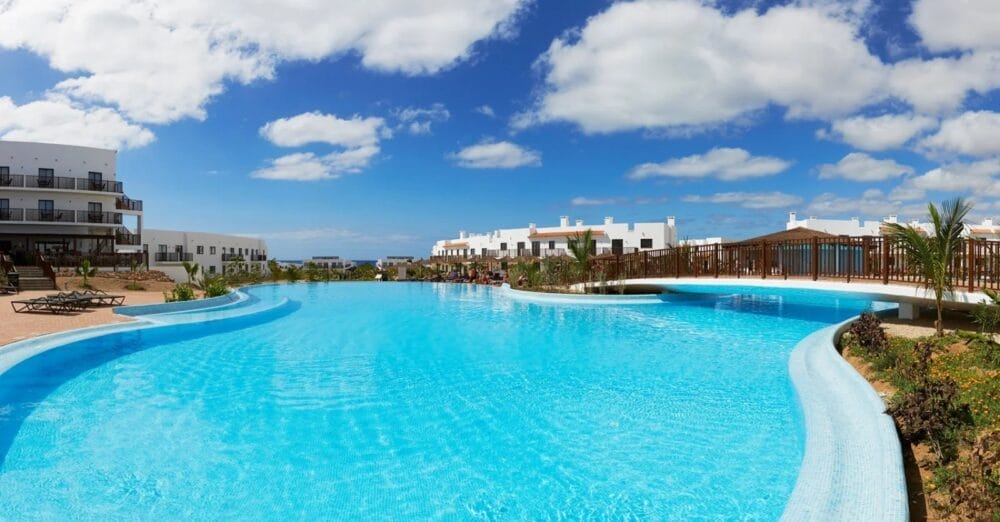 L'ampia e sinuosa piscina all'aperto del MELIA DUNAS BEACH RESORT è circondata da moderni edifici bianchi, sedie a sdraio e una passerella in legno, il tutto sotto un cielo blu brillante con nuvole sparse.