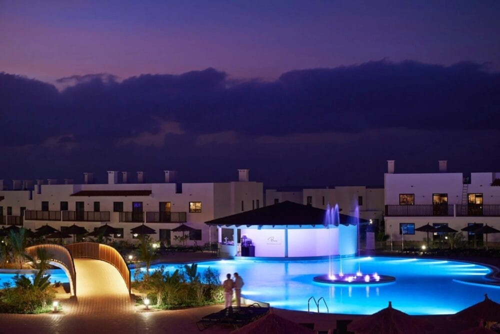 Una grande piscina illuminata risplende di blu al tramonto al MELIA DUNAS BEACH RESORT, circondato da moderni edifici bianchi. Una fontana illuminata decora il centro della piscina mentre due persone sono in piedi vicino al bordo, sotto profonde nuvole viola.