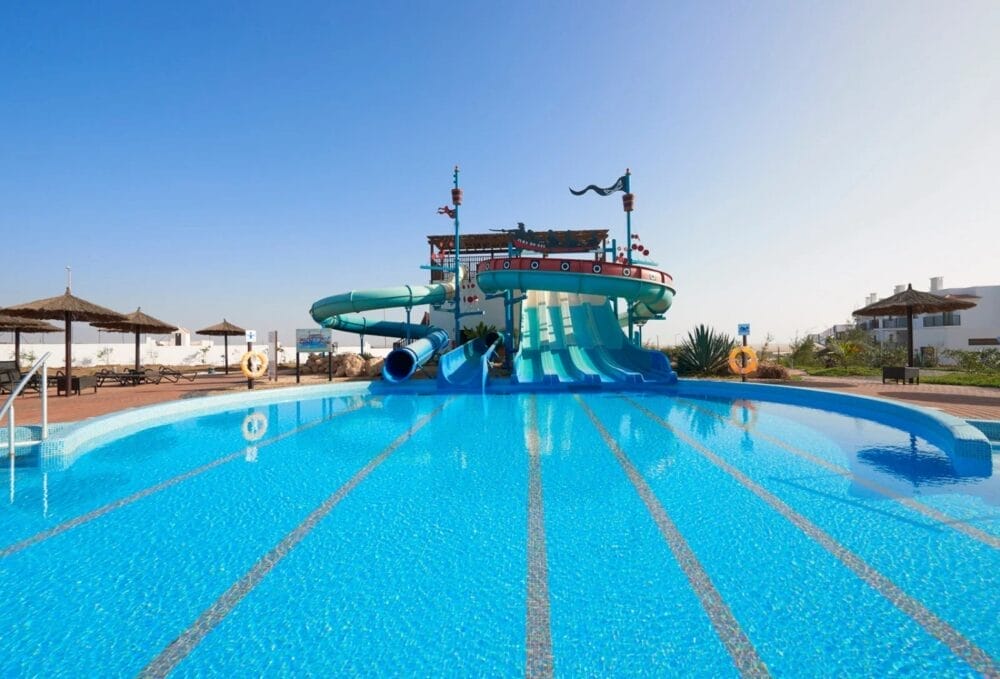 Una piscina all'aperto di colore azzurro chiaro con scivoli larghi e curvilinei e decorazioni di navi pirata al MELIA DUNAS BEACH RESORT, circondata da sedie a sdraio e ombrelloni di paglia sotto un cielo soleggiato.