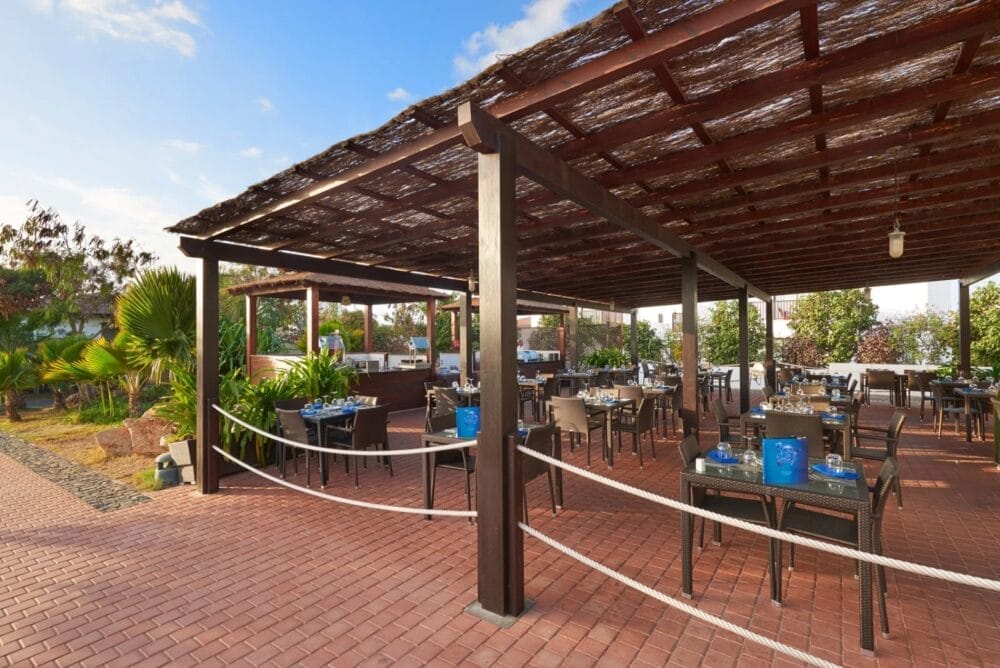 Ristorante all'aperto del Melia Dunas Beach Resort con pergola in legno scuro, pavimento in mattoni e tavoli e sedie ordinati. Circondato da piante e alberi verdi sotto un cielo azzurro, è il luogo perfetto per cenare godendosi l'atmosfera del resort sulla spiaggia.