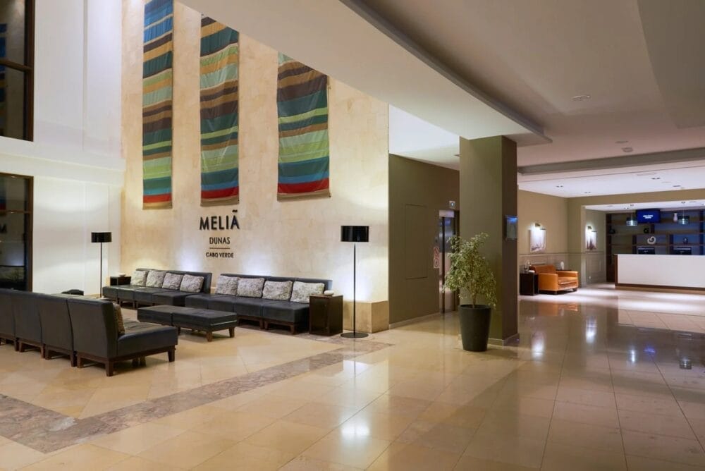 Moderna hall dell'hotel MELIA DUNAS Beach Resort con pavimento in piastrelle, divani neri e strisce di tessuto colorato sopra l'insegna "Meliá Dunas". Il banco della reception è visibile sullo sfondo a destra.