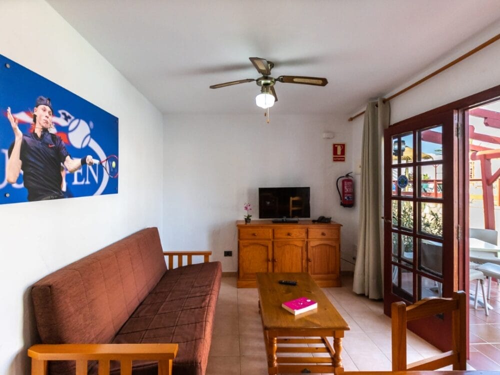 L'accogliente soggiorno del Sand Castillo Apartments presenta un futon marrone, mobili in legno, una TV, un ventilatore a soffitto e una grande porta a vetri. Un poster a tema tennistico è appeso alla parete e un libro è appoggiato sul tavolino.