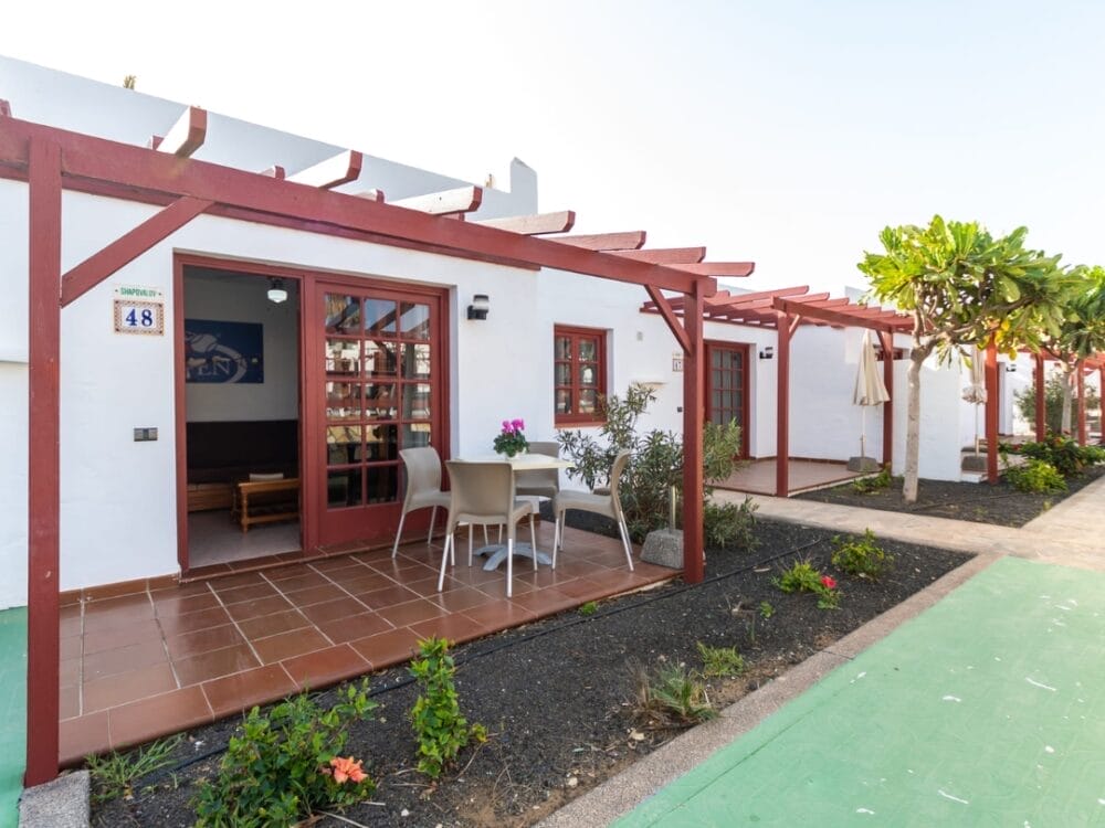 Un bungalow bianco del Sand Castillo Apartments presenta un pergolato in legno rosso, porte scorrevoli in vetro, un piccolo patio con tavolo e sedie e un giardino con piante e un albero lungo un vialetto pavimentato.