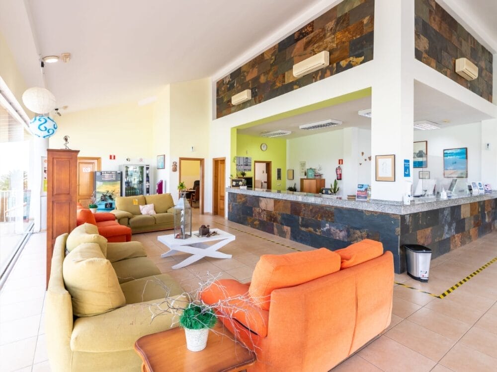 La luminosa hall dell'hotel Sand Castillo Apartments è caratterizzata da divani colorati, un tavolino bianco, un banco della reception con accenti in pietra, vivaci opere d'arte alle pareti, piante in vaso e grandi finestre che lasciano entrare la luce naturale.