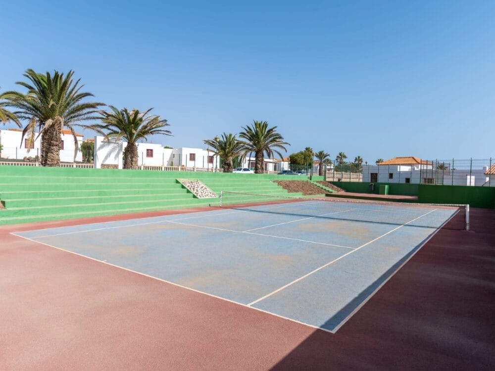 Campo da tennis all'aperto con superficie blu e rossa sbiadita, circondato da gradinate verdi e palme, sotto un cielo azzurro e limpido con gli appartamenti Sand Castillo e gli edifici bianchi sullo sfondo.