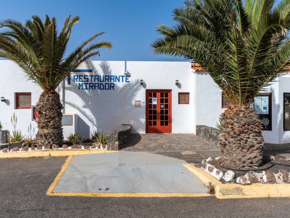 L'ingresso del Restaurante Mirador, vicino agli appartamenti Sand Castillo, è caratterizzato da un edificio bianco con una porta rossa, due grandi palme, un sentiero in pietra e un'insegna blu sopra la porta. Il cielo è limpido e soleggiato.