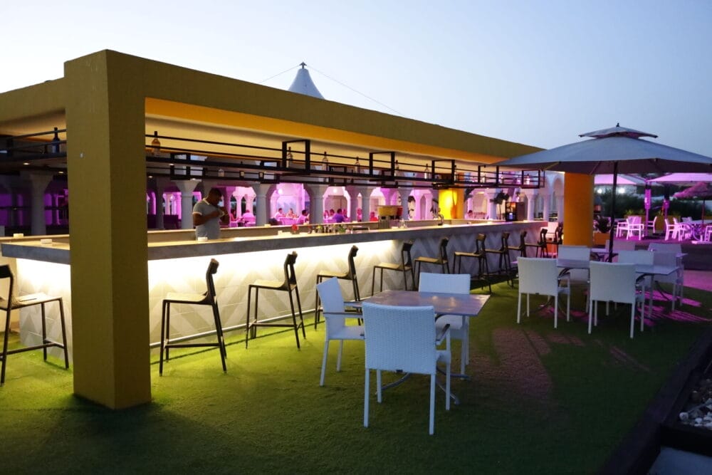 Bar all'aperto del MIRAGE HAMMAMET Resort con struttura gialla, sgabelli alti e tavoli e sedie bianchi su erba artificiale. Luci bianche e viola illuminano lo spazio vivace, dove gli ospiti si mescolano vicino al bar e sotto gli ombrelloni sullo sfondo.