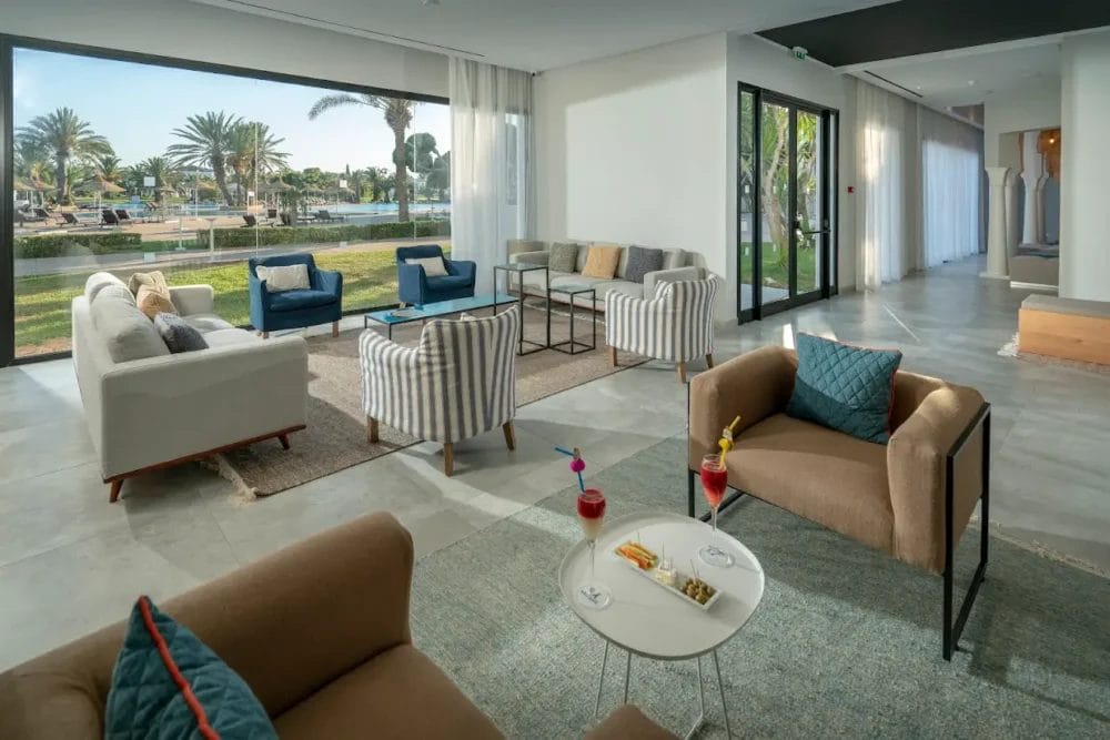 Moderna lounge del Mirage Hammamet Resort con poltrone, divani e tavolini, con bevande e snack. Le grandi finestre mostrano le palme, il prato verde e l'area della piscina. Interni luminosi, spaziosi e invitanti di un resort a 5 stelle.