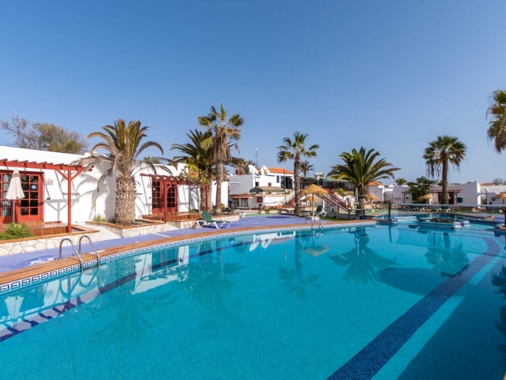 La soleggiata piscina all'aperto del Sand Castillo Apartments è circondata da palme e ville bianche con rifiniture rosse, con sedie a sdraio e un limpido cielo blu in alto.