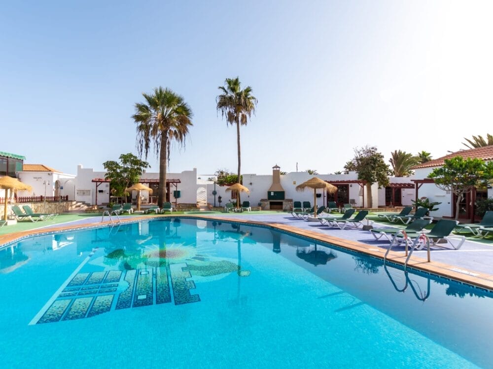 Piscina all'aperto con acqua cristallina, circondata da sedie a sdraio e ombrelloni verdi, con palme e bungalow bianchi degli Appartamenti Sand Castillo sullo sfondo, sotto un cielo limpido.