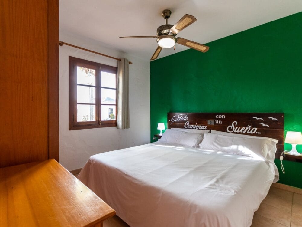 Un'accogliente camera degli Appartamenti Sand Castillo con un letto matrimoniale, biancheria da letto bianca, lampade su ogni lato, una parete verde d'accento, una testata in legno con testo spagnolo, un comò, una finestra e un ventilatore a soffitto.