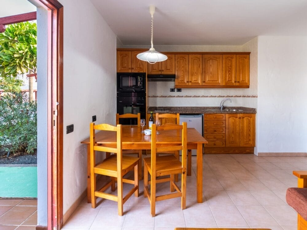 L'accogliente cucina degli APPARTAMENTI SAND CASTILLO è dotata di armadietti in legno e di un tavolo da pranzo abbinato a quattro sedie con tazze. Una luce a sospensione è appesa in alto e una porta aperta rivela una vegetazione lussureggiante oltre il pavimento piastrellato.
