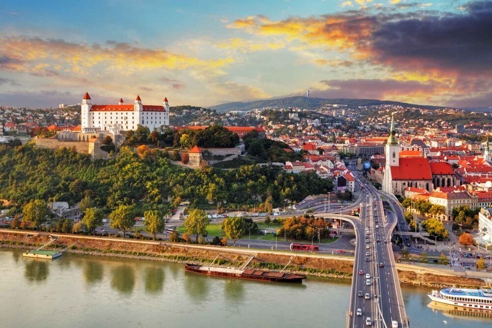 Una vista panoramica di Bratislava, in Slovacchia, con il Castello di Bratislava su una collina, il fiume e i ponti in primo piano, gli edifici colorati e la guglia di una chiesa sotto un drammatico cielo al tramonto: una tappa indimenticabile del vostro tour in pullman da Praga a Budapest.