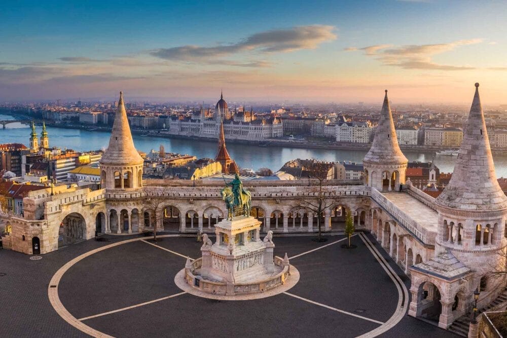 Vista aerea del Bastione dei Pescatori a Budapest, in Ungheria, con le sue torrette e la statua centrale. Il fiume Danubio e l'edificio del Parlamento ungherese risplendono al tramonto: un'occasione imperdibile per un tour Granturismo.