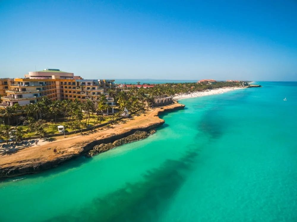 Veduta aerea della spiaggia tropicale di Varadero con acqua turchese, litorale sabbioso, palme e l'imponente complesso alberghiero MELIA VARADERO, sullo sfondo di un cielo azzurro e limpido.