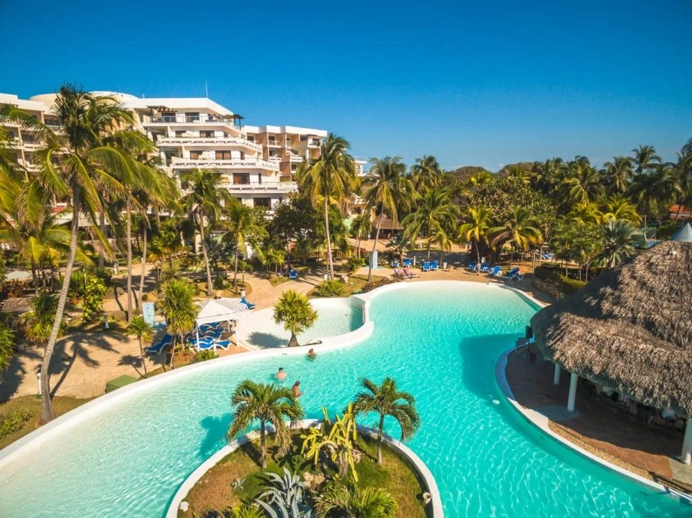 Un resort con un'ampia e sinuosa piscina all'aperto circondata da palme ed edifici con tetti di paglia, con un cielo azzurro e l'iconico hotel a più piani Melia Varadero sullo sfondo.