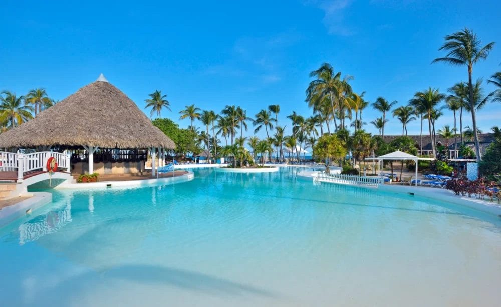 Una grande piscina tropicale con acqua cristallina al Melia Varadero, circondata da palme, sedie a sdraio e un bar con tetto di paglia, sotto un cielo blu brillante: la vostra fuga perfetta da Cuba.