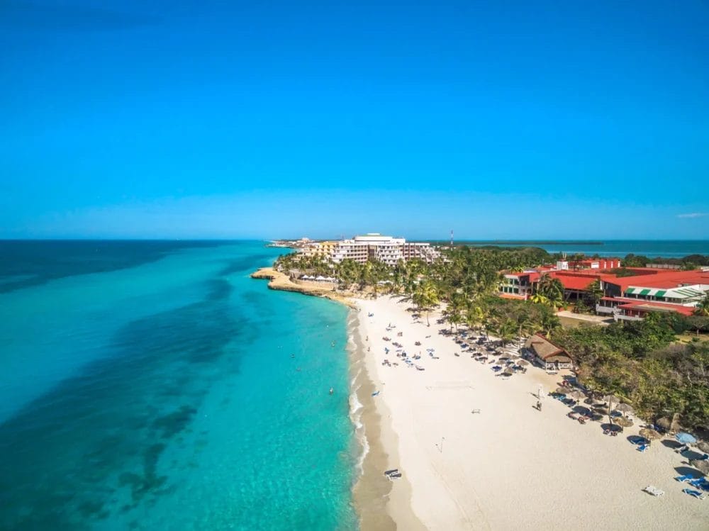 Vista aerea del Melia Varadero, uno splendido resort di Cuba con acqua turchese, sabbia bianca, lettini con ombrelloni, palme ed edifici del resort sotto un cielo azzurro e limpido.