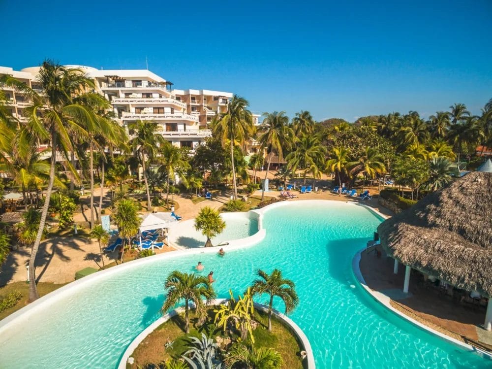 Una grande piscina si snoda tra le palme di questo resort di Cuba, con persone che si rilassano nell'acqua e sulle sedie a sdraio. Un hotel a più piani di Varadero e una capanna con il tetto di paglia sono visibili sotto un cielo azzurro e limpido.
