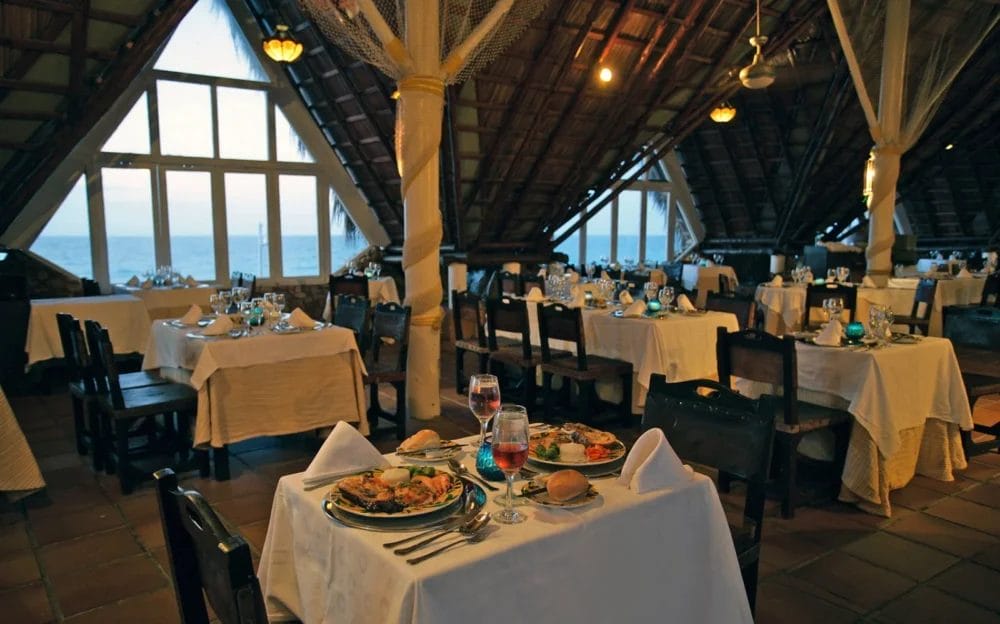L'accogliente ristorante all'aperto del Melia Varadero è caratterizzato da tavoli in legno apparecchiati per la cena con piatti e bevande deliziosi. Il tetto di paglia, le grandi finestre con vista sull'oceano e l'illuminazione calda catturano il fascino dei resort di Cuba.