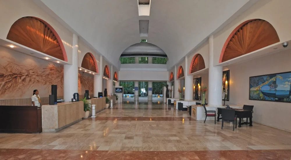 Ampia hall dell'hotel con pavimenti in marmo, alto soffitto ad arco, banchi della reception su entrambi i lati, una persona ad un banco, grandi opere d'arte a parete e porte di vetro che conducono all'esterno - evocando l'eleganza di un hotel di Varadero come il Melia Varadero.