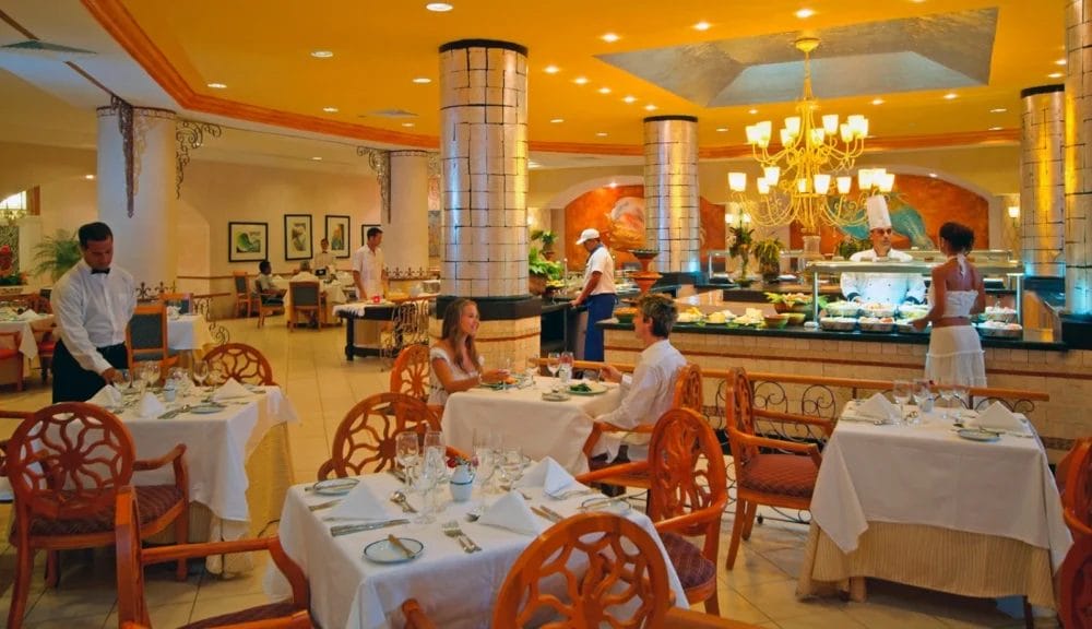 Elegante interno del ristorante dell'hotel MELIA VARADERO con luci calde, colonne decorate e un'area buffet. I commensali siedono ai tavoli vestiti di bianco mentre i camerieri in uniforme bianca servono il cibo. L'ambiente è accogliente e sofisticato a Varadero.