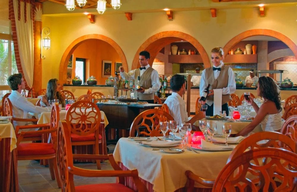 Un ristorante VARADERO illuminato da una luce calda e dall'arredamento elegante, dove camerieri in uniforme servono vino a commensali ben vestiti su tavoli MELIA apparecchiati con panni, bicchieri e piatti bianchi. Sullo sfondo, nicchie ad arco e ceramiche decorative.