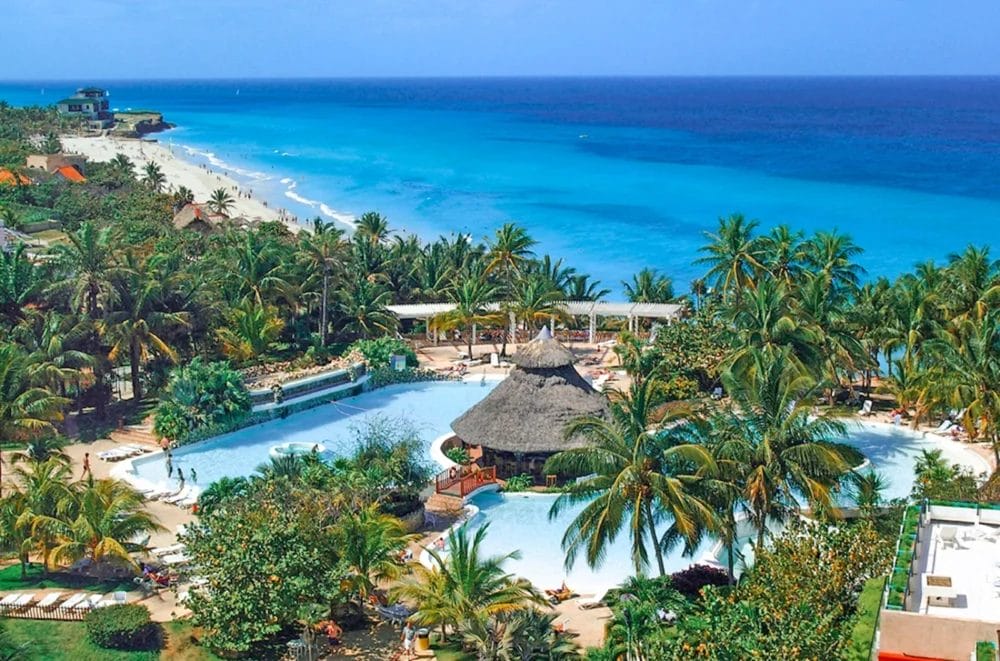 Un resort tropicale a Cuba, in riva all'oceano blu brillante, con palme, una grande piscina con bar sul tetto di paglia, lettini e giardini lussureggianti sotto un cielo limpido. La spiaggia sabbiosa si estende in lontananza al Melia Varadero.