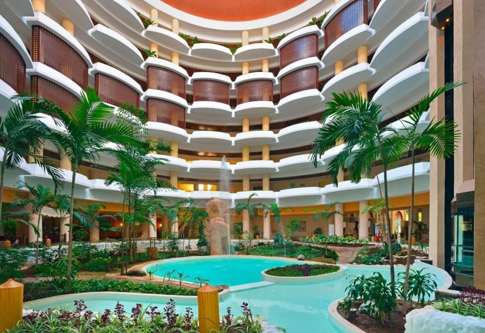 Uno spazioso atrio MELIA VARADERO a più piani e balconi, con piante tropicali, palme e una piscina interna turchese con una piccola fontana al centro, il tutto sotto un alto soffitto circolare di questo iconico resort di Cuba.