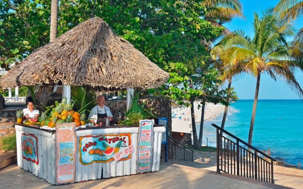 Un chiosco di succhi di frutta tropicale con un tetto di paglia si trova accanto alla spiaggia di un resort di Cuba. Due venditori sorridono dietro un bancone decorato con frutta e un'insegna colorata. Le palme e l'oceano turchese fanno pensare al vicino hotel Melia Varadero.