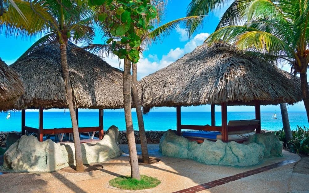 Cabine con tetto di paglia e letti sotto le palme si affacciano su un oceano tropicale blu brillante al MELIA VARADERO, uno splendido resort di Cuba, con cielo limpido e barche a vela visibili in lontananza.