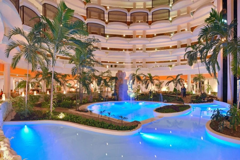 Atrio interno del Melia Varadero con diversi piani di balconi che si affacciano su una piscina sinuosa, piante tropicali, palme e una fontana d'acqua centrale, il tutto illuminato per creare un'atmosfera da resort.