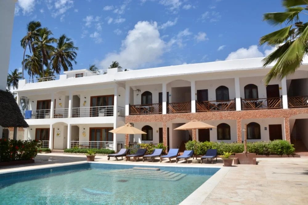 Un moderno edificio bianco a due piani con balconi si affaccia su una piscina circondata da sedie a sdraio e ombrelloni al Blu Marlin Beach Resort, con palme e cielo azzurro sullo sfondo.
