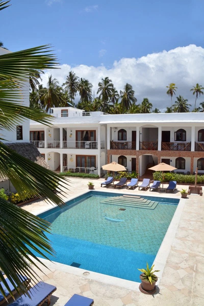 Una piscina trasparente circondata da sedie a sdraio e ombrelloni si trova nel cortile del Blu Marlin Beach Resort, un hotel bianco a due piani con balconi, palme e un cielo azzurro sullo sfondo.