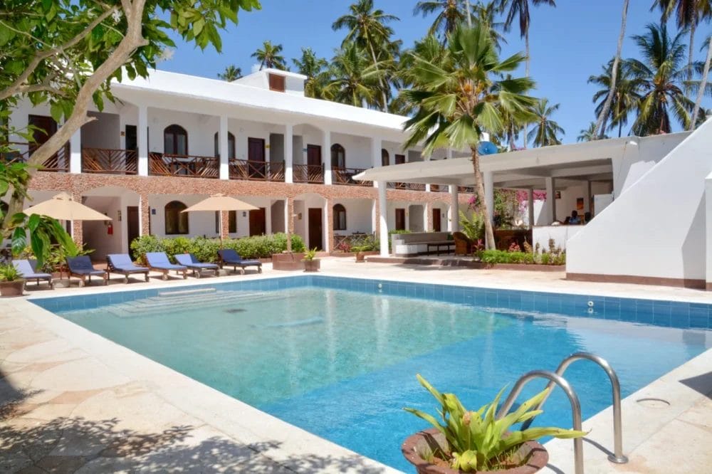 Un resort tropicale sulla spiaggia con edifici bianchi a due piani, balconi e una piscina blu chiaro circondata da sedie a sdraio, ombrelloni, palme e verde lussureggiante sotto un cielo soleggiato al BLU MARLIN BEACH RESORT.