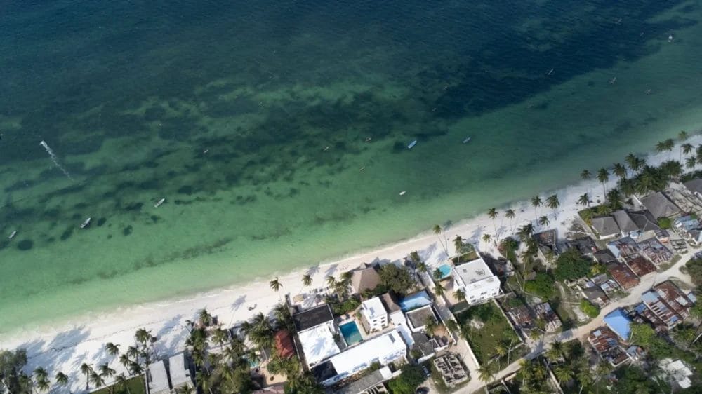Veduta aerea di Marlin Beach con sabbia bianca, acqua turchese e cristallina, palme ed edifici costieri lungo la riva. Piccole imbarcazioni e bagnanti sono visibili nel mare poco profondo vicino al BLU MARLIN BEACH RESORT.