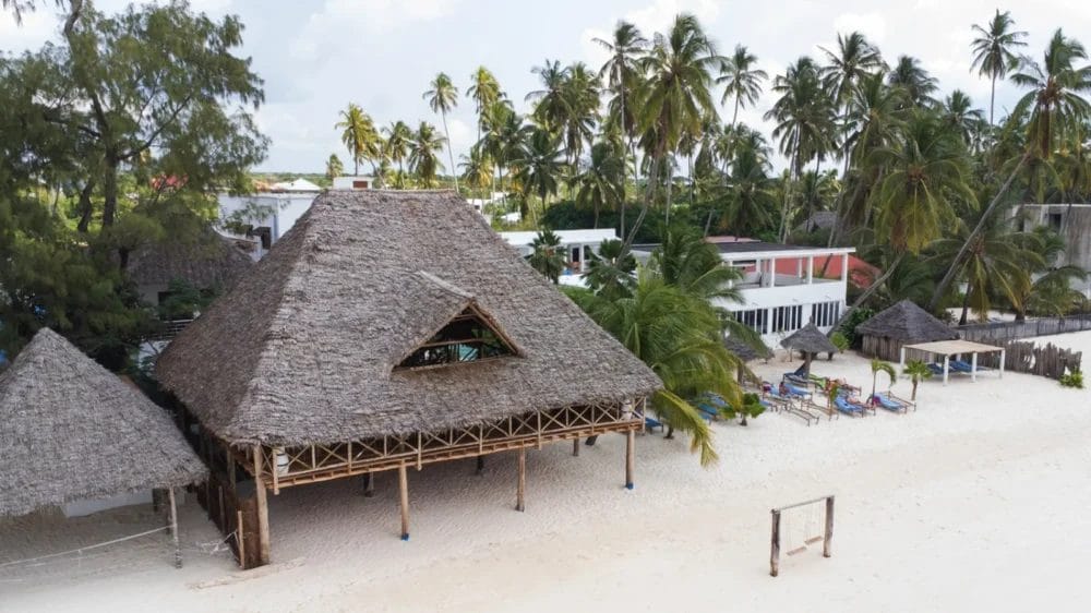 Un grande edificio con tetto di paglia sulla spiaggia di Marlin, circondato da palme e sedie a sdraio. Edifici moderni e verde lussureggiante sono visibili sullo sfondo sotto un cielo parzialmente nuvoloso al BLU MARLIN BEACH RESORT.