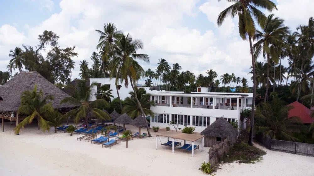 Un hotel bianco sulla spiaggia di Marlin Beach, circondato da alte palme, offre sedie a sdraio blu sull'arenile e strutture con tetti di paglia nelle vicinanze, sotto un cielo parzialmente nuvoloso: una perfetta fuga da una località balneare.