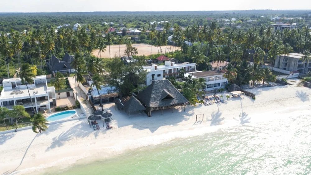 Veduta aerea del BLU MARLIN BEACH RESORT con palme, spiaggia di sabbia bianca, acqua limpida e turchese, piscina e diversi edifici circondati dal verde. Le case e la vegetazione si estendono sullo sfondo.