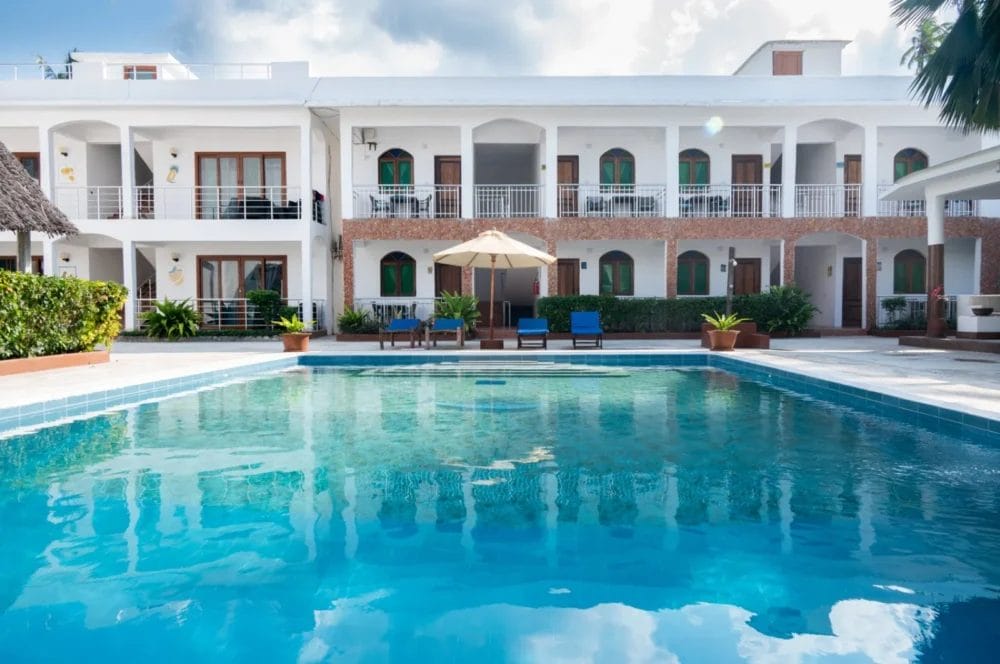 Una piscina azzurra e trasparente si trova di fronte all'edificio bianco a due piani del BLU MARLIN Beach Resort, con porte ad arco, balconi, sedie da patio, piante in vaso e un ombrellone sotto un cielo parzialmente nuvoloso.