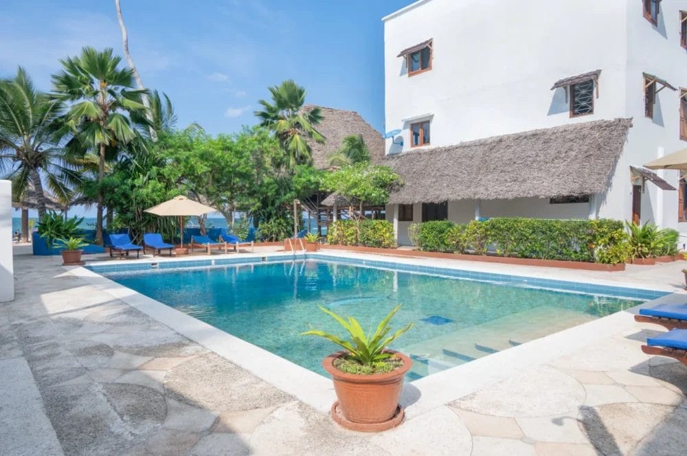 La serena piscina all'aperto del BLU MARLIN BEACH RESORT, circondata da sedie a sdraio blu, ombrelloni, palme e un edificio bianco con finestre in legno e tetto di paglia, sullo sfondo del cielo e dell'oceano blu di Marlin Beach.