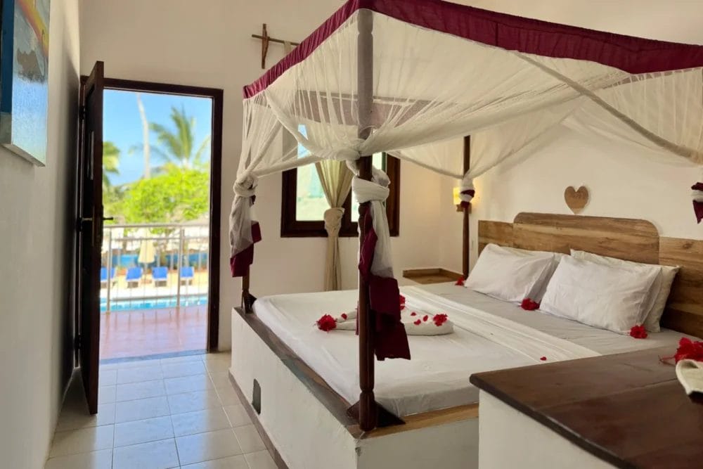 L'accogliente camera da letto del BLU MARLIN BEACH RESORT presenta un letto a baldacchino in legno avvolto in una zanzariera bianca e decorato con fiori rossi. Pareti bianche, pavimenti piastrellati e una porta aperta rivelano un balcone con vista sulle palme e sul cielo blu.