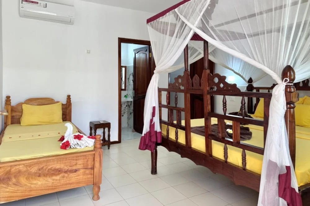 La luminosa camera del BLU MARLIN BEACH RESORT dispone di un letto singolo in legno e di un letto matrimoniale a baldacchino, entrambi con lenzuola gialle. Gli asciugamani a forma di cigno con fiori sono appoggiati sul letto singolo e una porta aperta conduce al bagno.