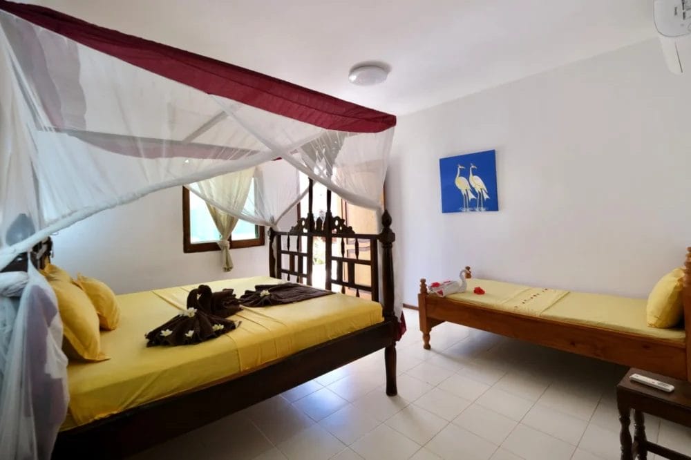 La luminosa camera da letto del BLU MARLIN BEACH RESORT presenta un letto matrimoniale e uno singolo in legno con biancheria da letto gialla. Il letto matrimoniale ha un baldacchino bianco. La luce naturale entra dalla finestra e un dipinto di due uccelli adorna la parete.