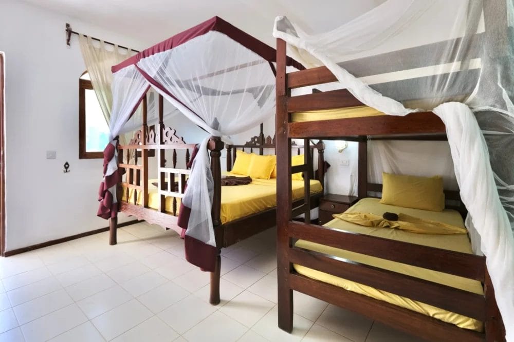 La luminosa camera da letto del BLU MARLIN BEACH RESORT, con pavimento in piastrelle bianche, presenta un letto a baldacchino in legno e un letto a castello in legno, entrambi con lenzuola gialle e zanzariere. La luce del sole entra da una finestra con tende leggere.