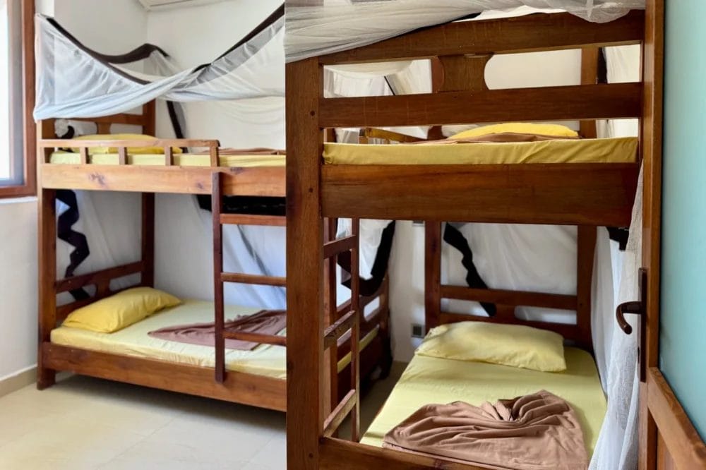 Una luminosa camera del BLU MARLIN BEACH RESORT presenta due letti a castello in legno con lenzuola gialle, coperte marroni piegate ordinatamente e zanzariere bianche drappeggiate sopra la testa. La luce del sole entra da una finestra, creando un accogliente rifugio a Marlin Beach.